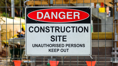 A 'Danger, Construction site, Unauthorised Persons Keep Out' sign mounted on a wire fence in front of a building site.